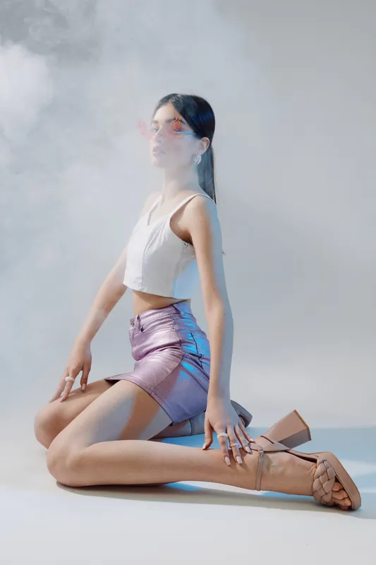 Young woman beauty fashion model kneels on floor wearing a white crop top and shiny purple skirt. Studio shot with soft lighting and smoke effect on a neutral background.