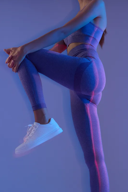 Woman sport fitness stretching in textured blue leggings and sports bra with white sneakers, preparing for exercise in soft neon lighting and moody studio setting.