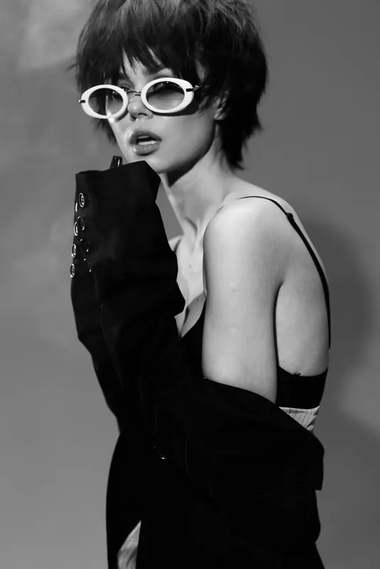 Black and white portrait of a stylish woman wearing white sunglasses and a black coat with detached expression