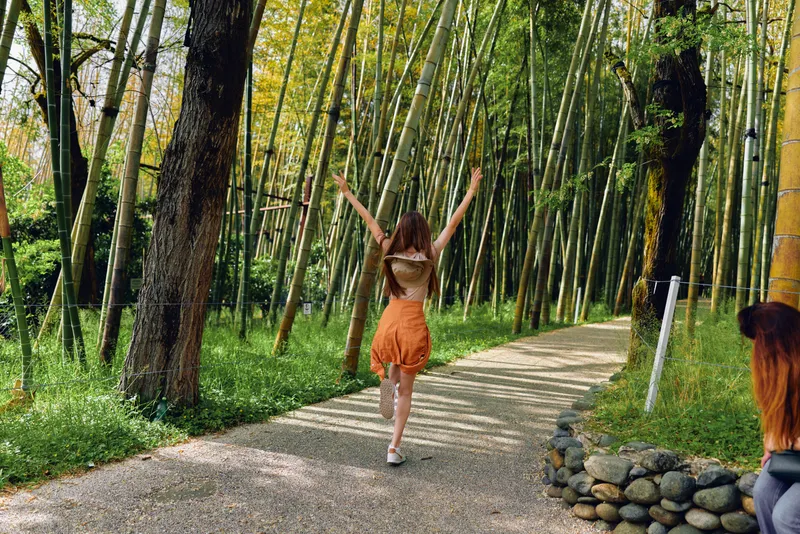 Woman walking back on a bamboo path through a sunlit forest, arms raised in carefree joy. Young traveler in orange skirt on a gravel trail surrounded by green trees and serenity