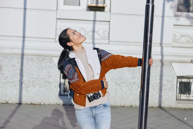Woman stretches toward a streetlamp in an urban setting, wearing a color block jacket and jeans, exploring balance and movement in daylight. She stands on a sidewalk beside a pale facade, exuding