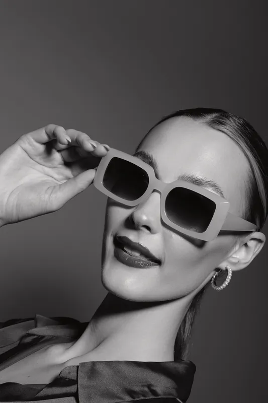 Elegant white woman with light skin wearing large sunglasses and hoop earrings poses confidently in black and white studio portrait.