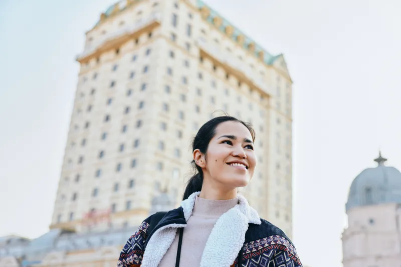A smiling woman outdoors in front of a tall city building, showcasing urban fashion with a cozy scarf and jacket, capturing a confident moment of travel, exploration and architectural backdrop.