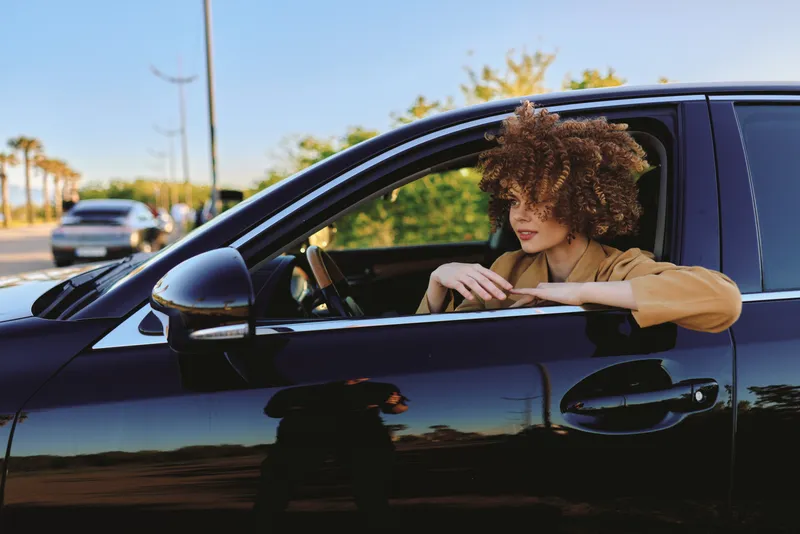 Curly haired woman leaning out of a sleek car window, enjoying a sunny street scene, city breeze in her hair. Retro film color evokes casual travel, freedom, and everyday moments.