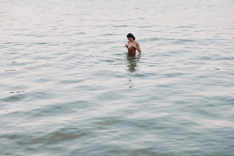 A lone figure stands waist-deep in clear sea water, wearing a swimsuit, with a peaceful stance and gentle waves, capturing a serene moment of solitude and reflection.