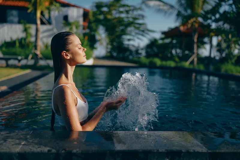 A poolside scene shows a woman in a white swimsuit enjoying relaxation, sunlight highlighting her profile as water splashes softly, conveying outdoor leisure and lifestyle calm