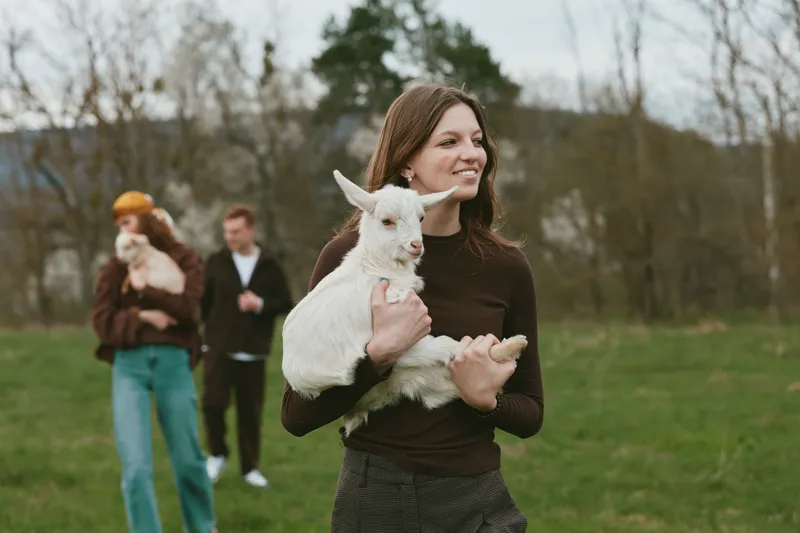 Young woman with long brown hair holds a white baby goat outdoors in a green field during an overcast day, smiling gently with two people blurred in the background.