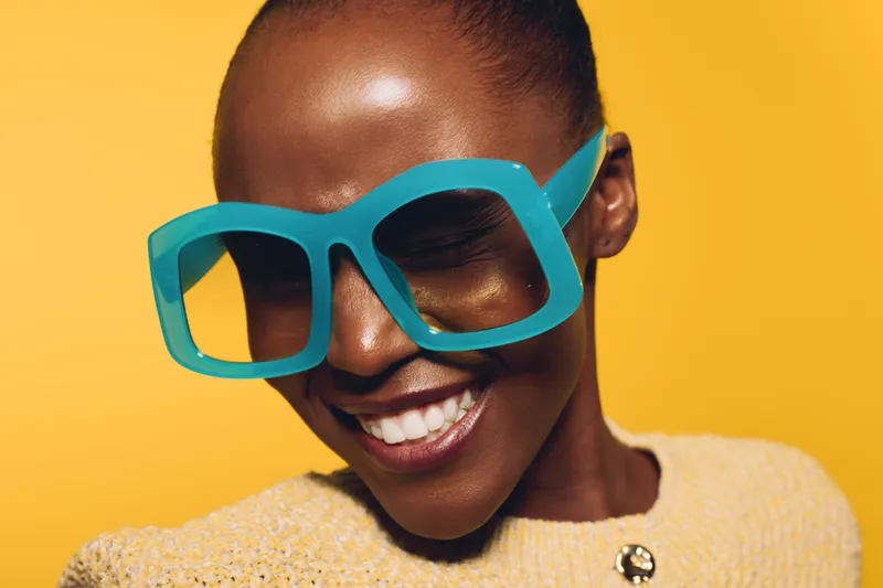 Smiling dark skinned African woman wears large turquoise sunglasses and beige sweater against yellow background. Close-up portrait with joyful mood and trendy fashion concept.