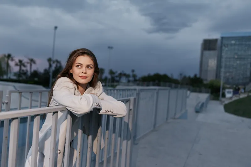 Young woman in white jacket leaning on fence outdoors with modern cityscape under cloudy sky at dusk, casual style, contemplative mood, urban lifestyle, fashion