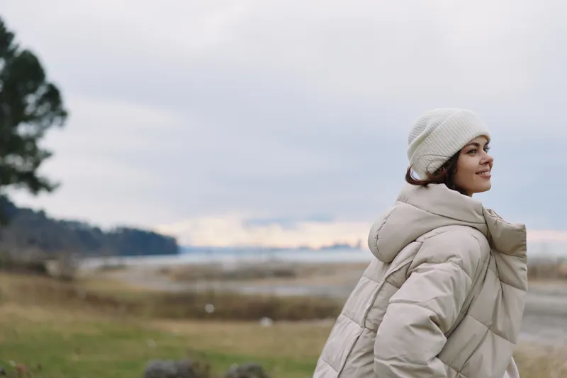 Woman in a warm beige winter coat and white beanie stands outdoors along a calm landscape with water and distant trees, serene mood, soft natural light.