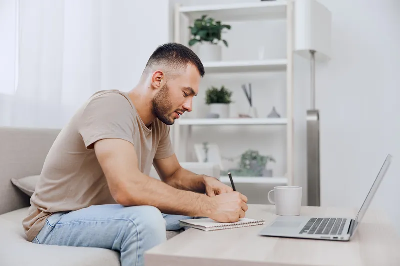 Focused man writing in notebook while sitting comfortably at home. The modern workspace features a laptop and a calming atmosphere, perfect for productivity.
