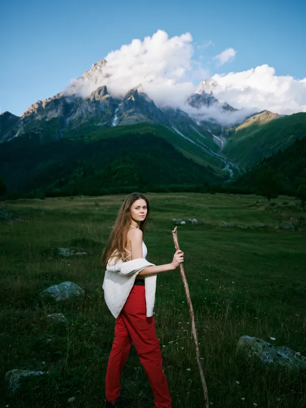 Woman hiking in a rugged meadow with a wooden staff, red pants and white shawl, set against towering mountains, blue sky and green valley, exploration and outdoor adventure