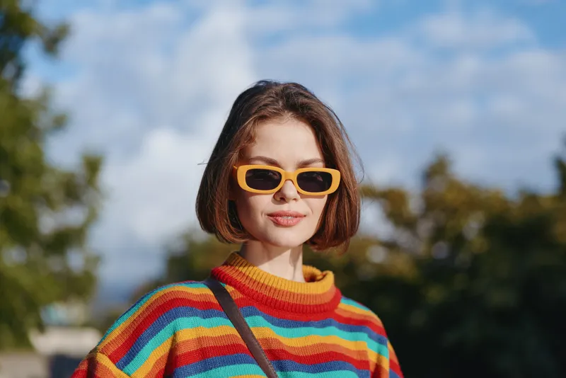 Young woman portrait wearing yellow sunglasses and rainbow sweater, short hair bob, smiling outdoors in an urban park, casual outfit and travel lifestyle fashion with sunny warm tones.