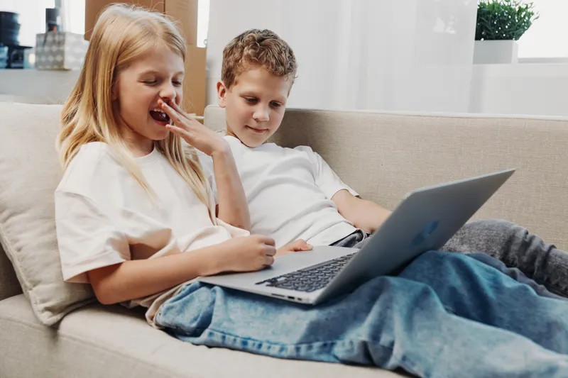 Two children sitting on a sofa using a laptop together, the girl yawns while the boy focuses on the screen. Cozy home interior with natural light from the window.