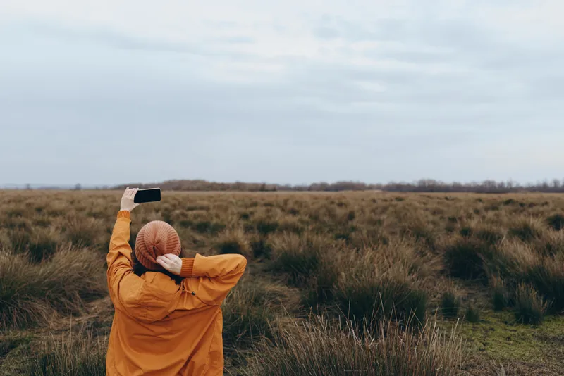 Woman outdoors lifestyle, standing in a grassy field with a warm jacket, holding a phone up, taking photo, hat near her head, cloudy sky, quiet nature, distant meadow horizon, soft wind