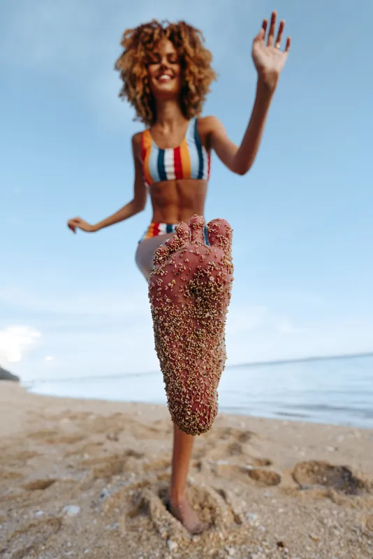 Smiling woman with curly hair in a colorful bikini stands on a sandy beach by the ocean, raising one hand and posing for a photo, summer travel lifestyle, natural light, joyful mood