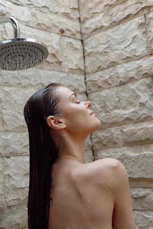 Woman enjoying outdoor shower with wet hair and closed eyes, surrounded by textured stone wall, natural light highlighting serene and relaxed mood under water droplets.
