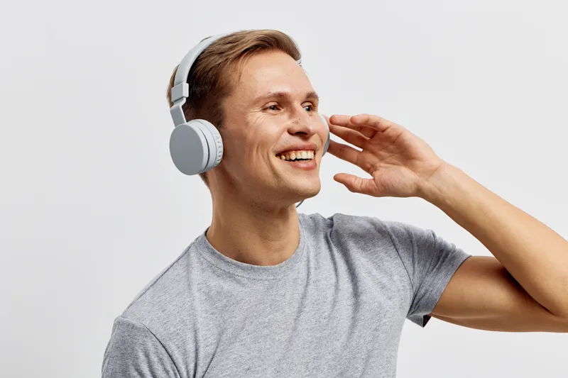 Close-up of cheerful young Caucasian man wearing gray t-shirt and large headphones, enjoying music with a bright smile, isolated on plain background, studio shot. Lifestyle and leisure concept.