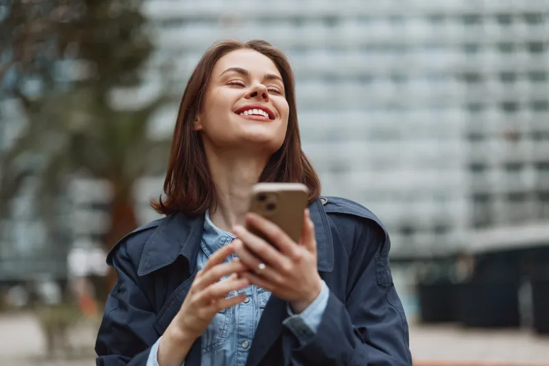 Young woman with long brunette hair smiles joyfully while holding a smartphone outdoors on a cool day in casual denim and dark jacket.