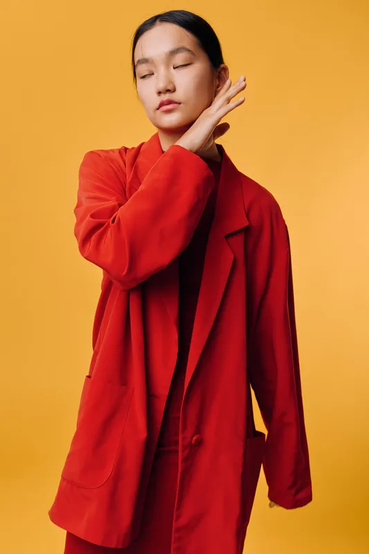 Young woman with closed eyes wearing oversized red blazer and matching outfit posing thoughtfully against orange background. Portrait of fashion model with sleek hairstyle.