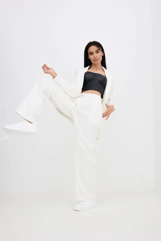 Young woman in white pants and blazer with black crop top posing dynamically against minimalistic white background, showcasing modern casual fashion style.
