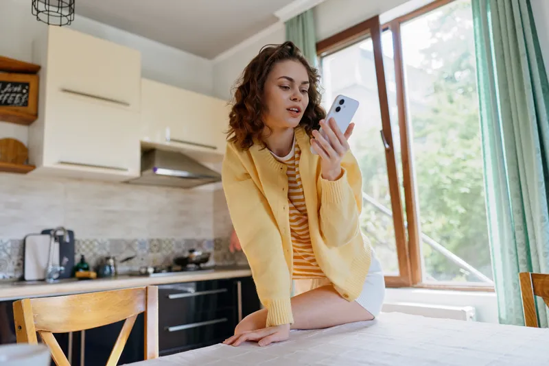 Young woman in a yellow cardigan sits at a kitchen island, scrolling on a smartphone with a curious relaxed smile.