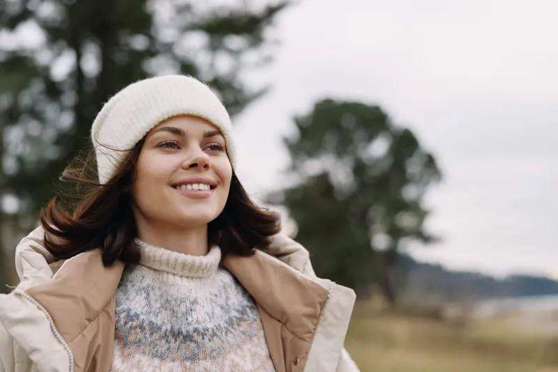 Woman enjoying outdoor winter weather in a warm beige coat, knit sweater and white beanie, smiling as a scenic landscape and trees frame the open countryside