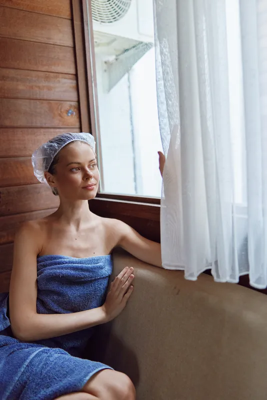 Relaxed woman wrapped in a blue towel with a shower cap, sitting by the window in a wooden room with soft natural light and sheer white curtains.