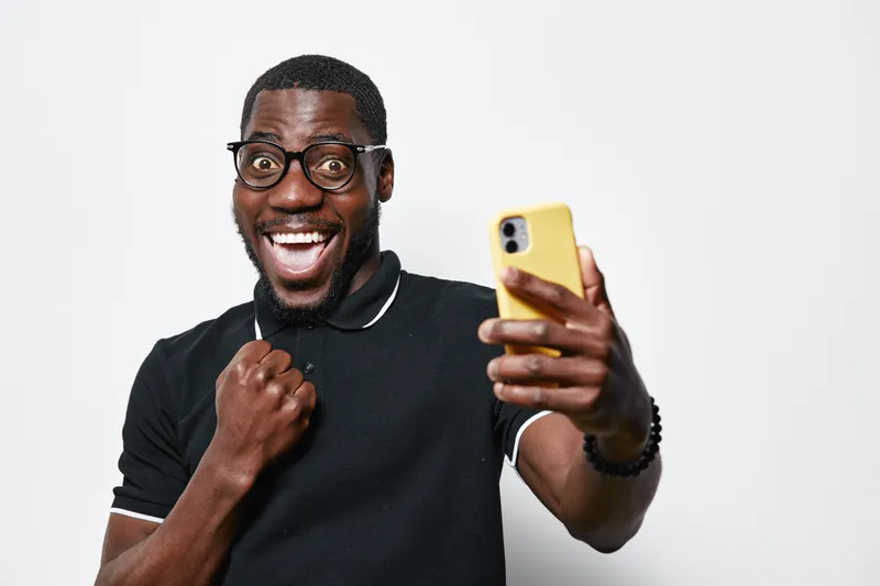 African American man with dark skin and glasses taking selfie with smartphone, smiling and showing excitement in casual black shirt on white background.