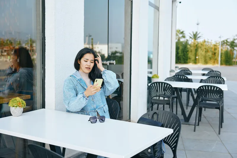 Woman seated at an outdoor cafe table using a phone, sunglasses on the table, casual attire, relaxed urban moment with glass storefront and daylight, a calm scene of everyday communication and