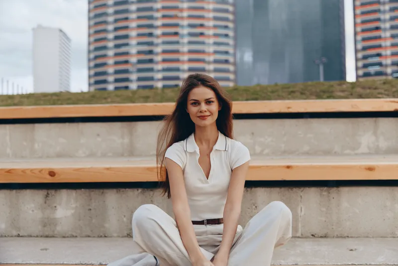 Woman sitting relaxed outdoors in casual white outfit with modern cityscape background, confident natural smile, urban fashion lifestyle spring concept