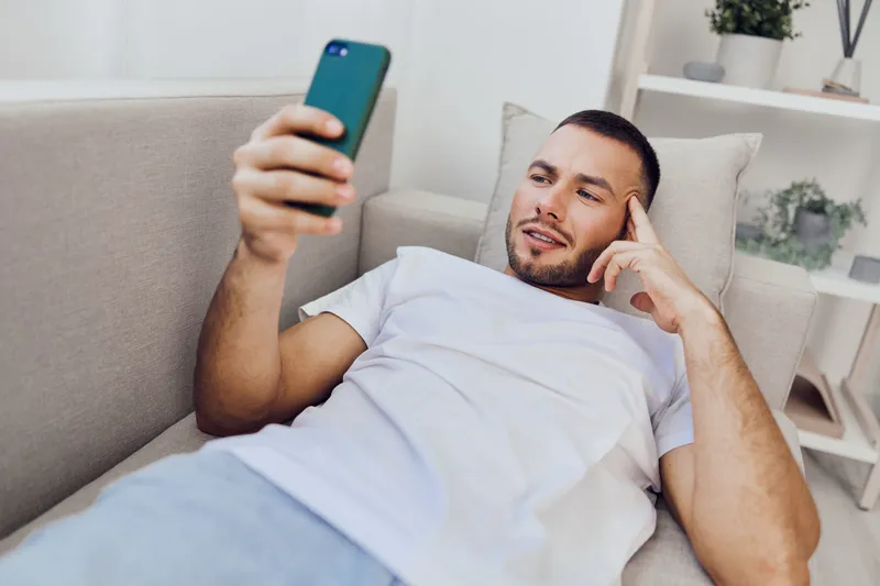 Relaxed man using smartphone while lying on a couch, exploring digital content with a thoughtful expression. Perfect for modern lifestyle and technology themes.