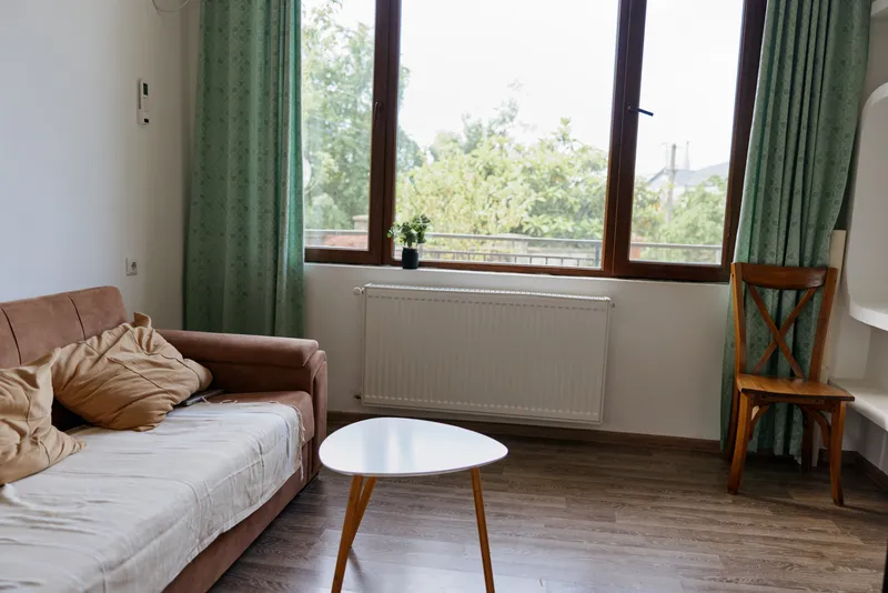 Cozy minimalist living room with a beige sofa, round white table, wooden chair, green curtains and a large window letting in soft daylight, creating a calm home atmosphere perfect for relaxing