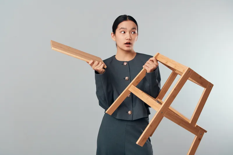 Surprised woman holding broken wooden chair frame and detached piece with worried expression on light background. Female in formal suit with dark hair in interior furniture repair or damage concept.