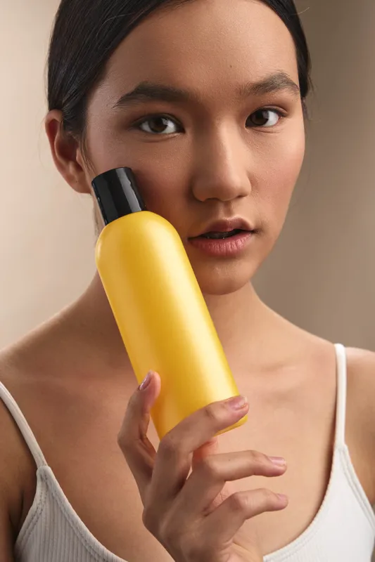 Asian woman holds a yellow cosmetic bottle close to her face, presenting a clean beauty product in a soft studio setting with gentle light and a calm, focused expression.