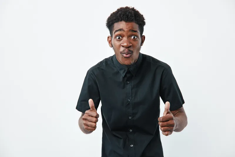 Young black man with dark skin wearing black shirt showing thumbs up gesture with surprised facial expression on white background. Positive, confident and casual style.