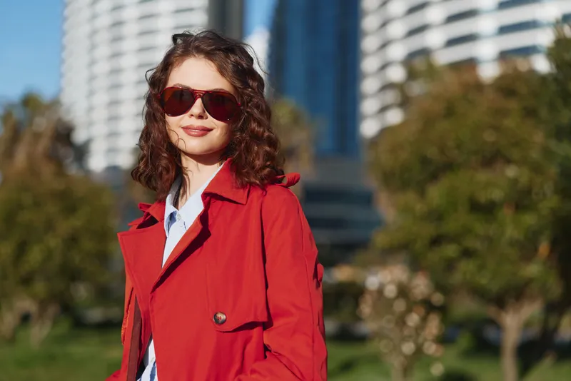 Young woman wearing a red coat and sunglasses stands confidently outdoors in an urban park on a sunny day, smiling softly as modern buildings rise behind lush green trees.