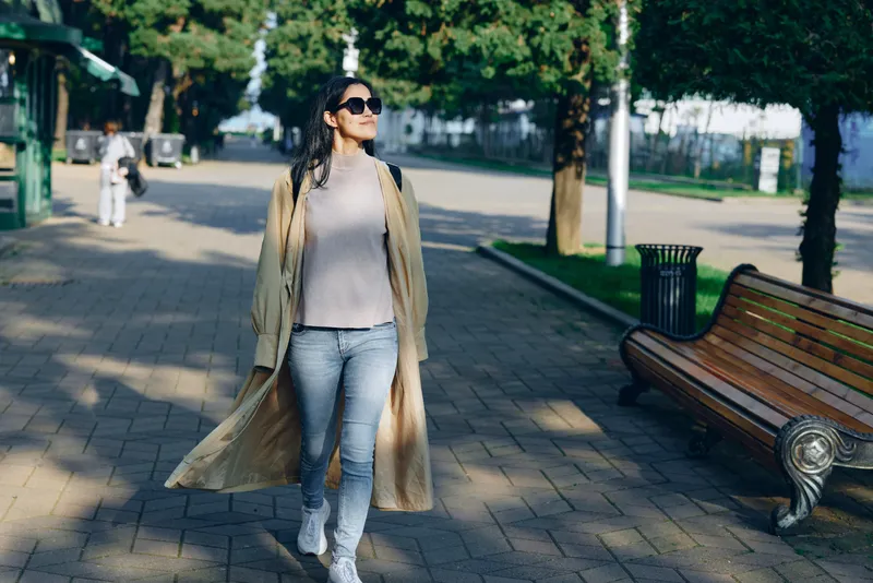 Woman walking in a sunlit park wearing a beige coat, light sweater and jeans with sunglasses, strolling along a paved path beside benches and trees surrounding