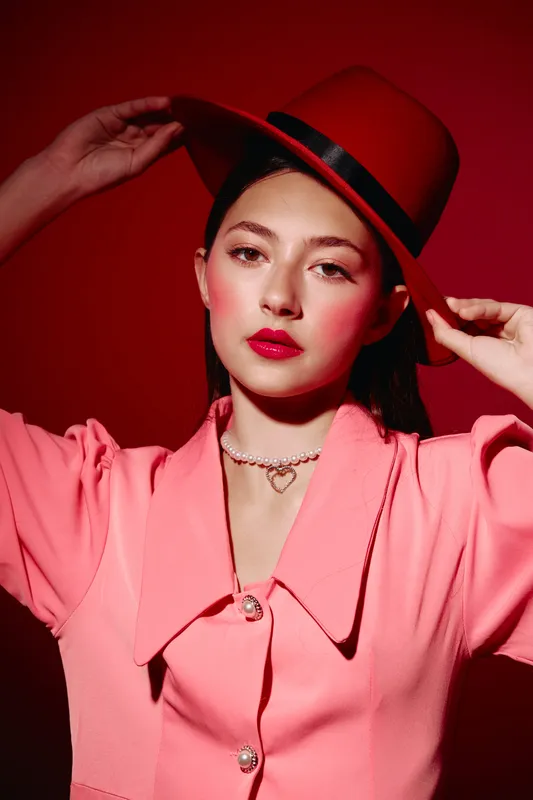 Young stylish woman wearing a bright pink coat and red hat poses against a deeply vivid red background. Her makeup highlights red lipstick and blush for a high fashion beauty look.
