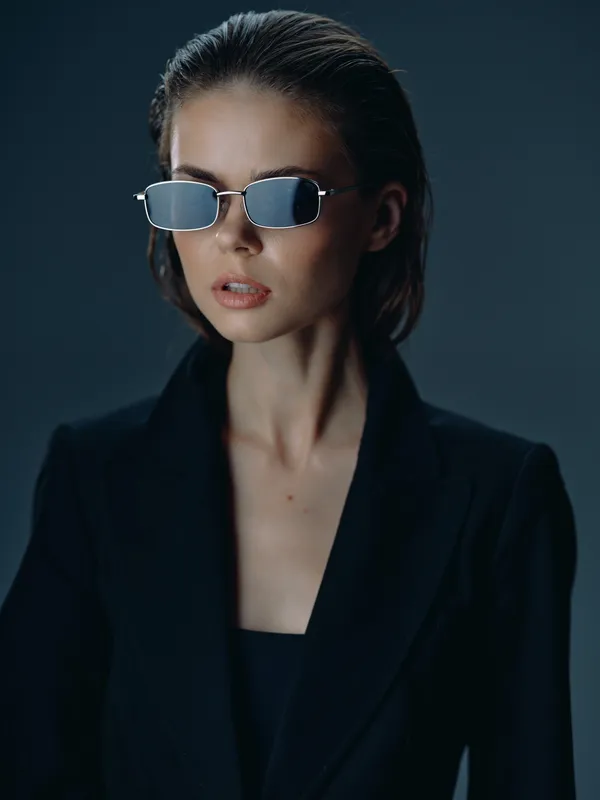 Stylish woman with sunglasses wearing a black blazer against dark background, showcasing modern fashion and confident expression.