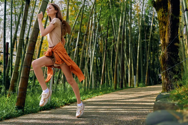 Woman jumping on bamboo forest path with backpack and orange jacket tied at waist, sneakers on feet, joyful hiking on winding trail with sunlight filtering through tall green grove.