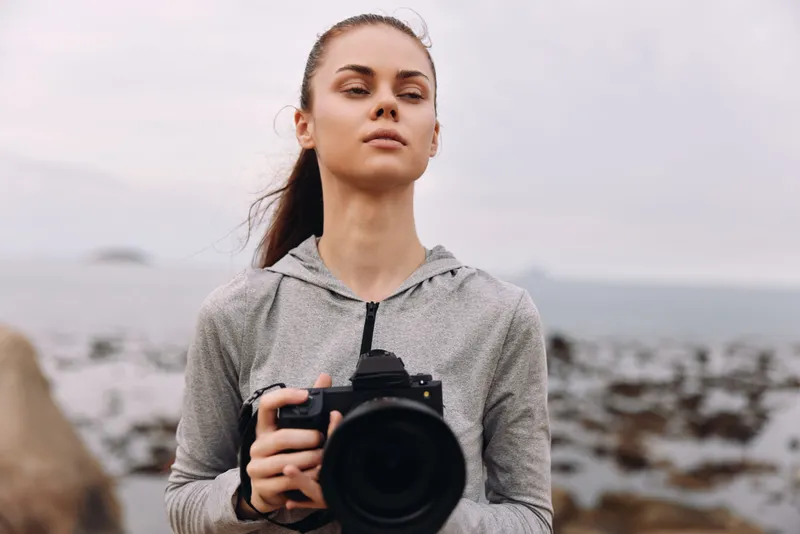 Female photographer on the beach holding a camera, dressed in casual sportswear, exuding confidence and focus with a moody ocean background
