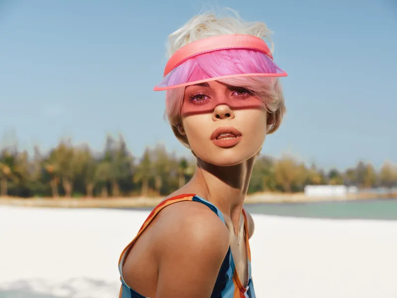 A fashionable woman on a sunny beach wears a pink translucent visor, colorful swimsuit, and windswept hair, creating a bold contemporary summer portrait under bright light.
