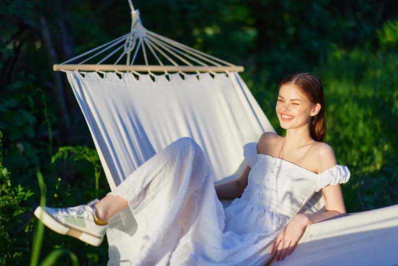 Young woman in a white dress relaxing in a hammock, smiling joyfully in a lush green garden during a sunny day Fresh outdoor atmosphere and summer vibes