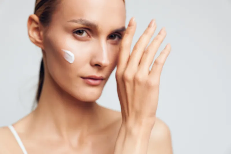 Young woman with clear skin applying white face cream on cheek, close-up on light background, skincare and beauty concept, natural makeup and self-care routine.