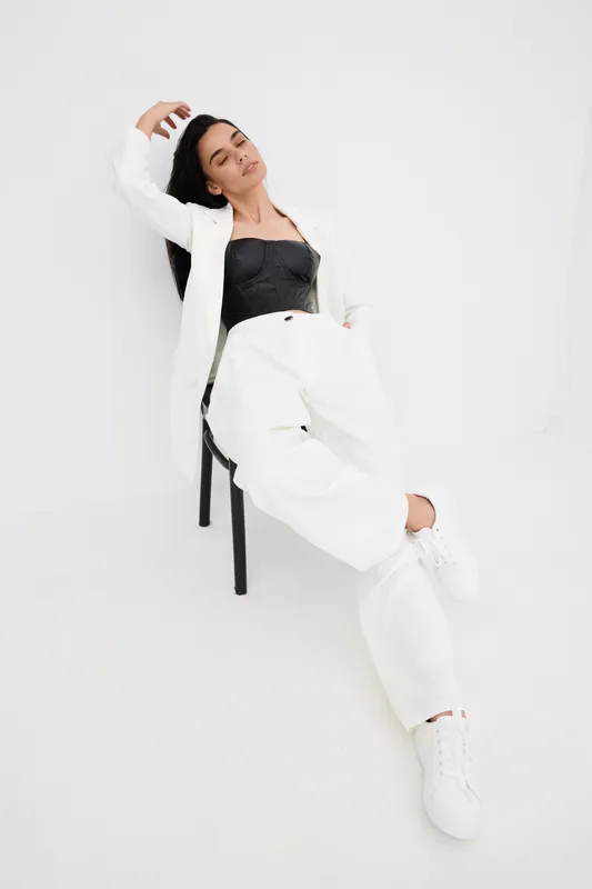Relaxed woman sitting on chair wearing white jacket and wide-leg pants with black crop top, casual fashion pose against minimalist white background.