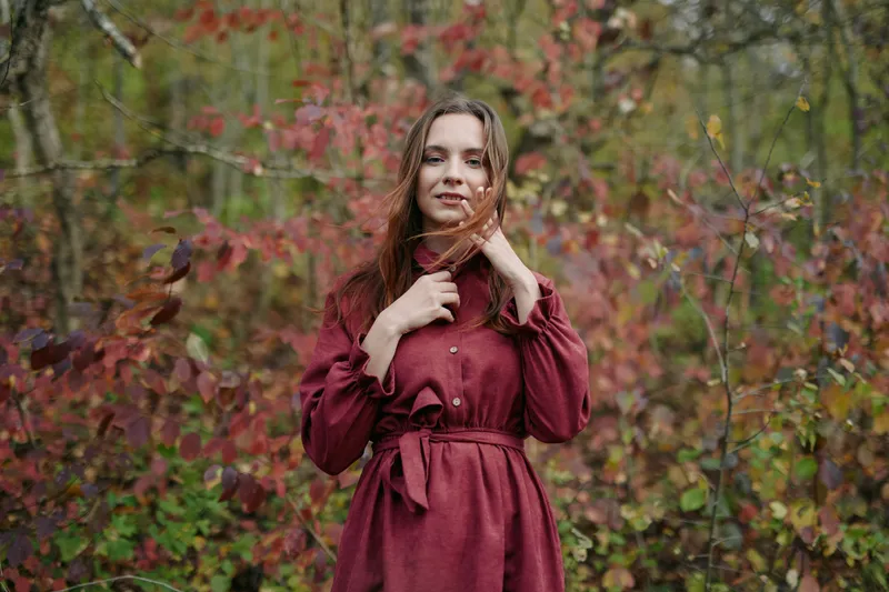 A candid portrait of a woman in a burgundy dress standing in a autumn forest, capturing authenticity and heartfelt expression amid falling leaves.