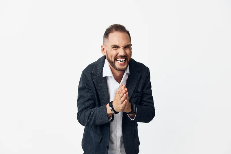 Professional man in business attire stands with hands clasped, smiling confidently. Isolated bright white background highlights focused emotion and attentive presence.
