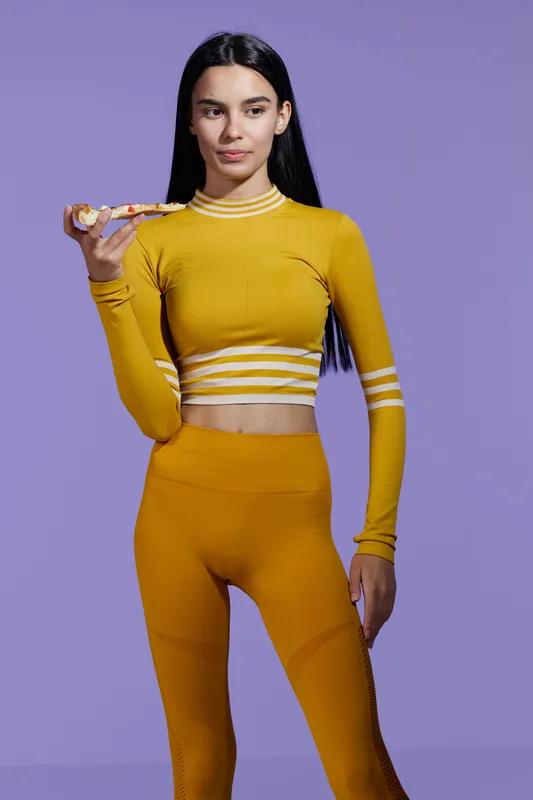 woman sport fitness in yellow activewear holding a healthy snack, standing against a purple background, young slender athletic female with long black hair, wellness and nutrition