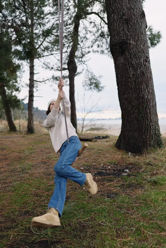 Person on a rope swing in a forested park beside a lake, casual outfit with jeans and boots, capturing a playful outdoor moment